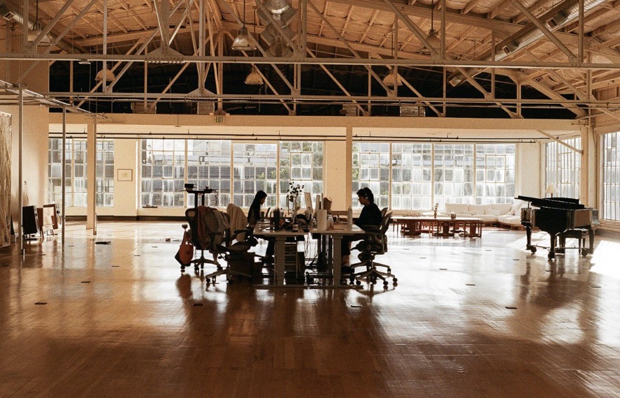 A quiet loft with tall arched windows, wood floors, two people working at a shared desk, and a piano in the corner.